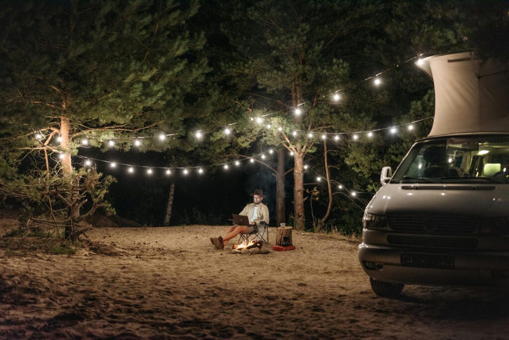 Man Sitting with a Laptop on a Beach near a Campervan and under String Lights at Night